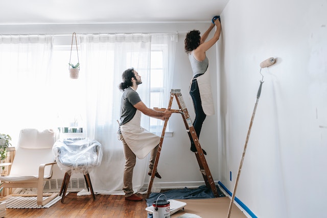 One person on a ladder painting a wall while another person holds the ladder